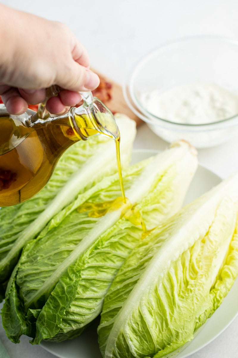 a hand pouring oil onto romaine lettuce heads with a bowl of blue cheese dressing in the background