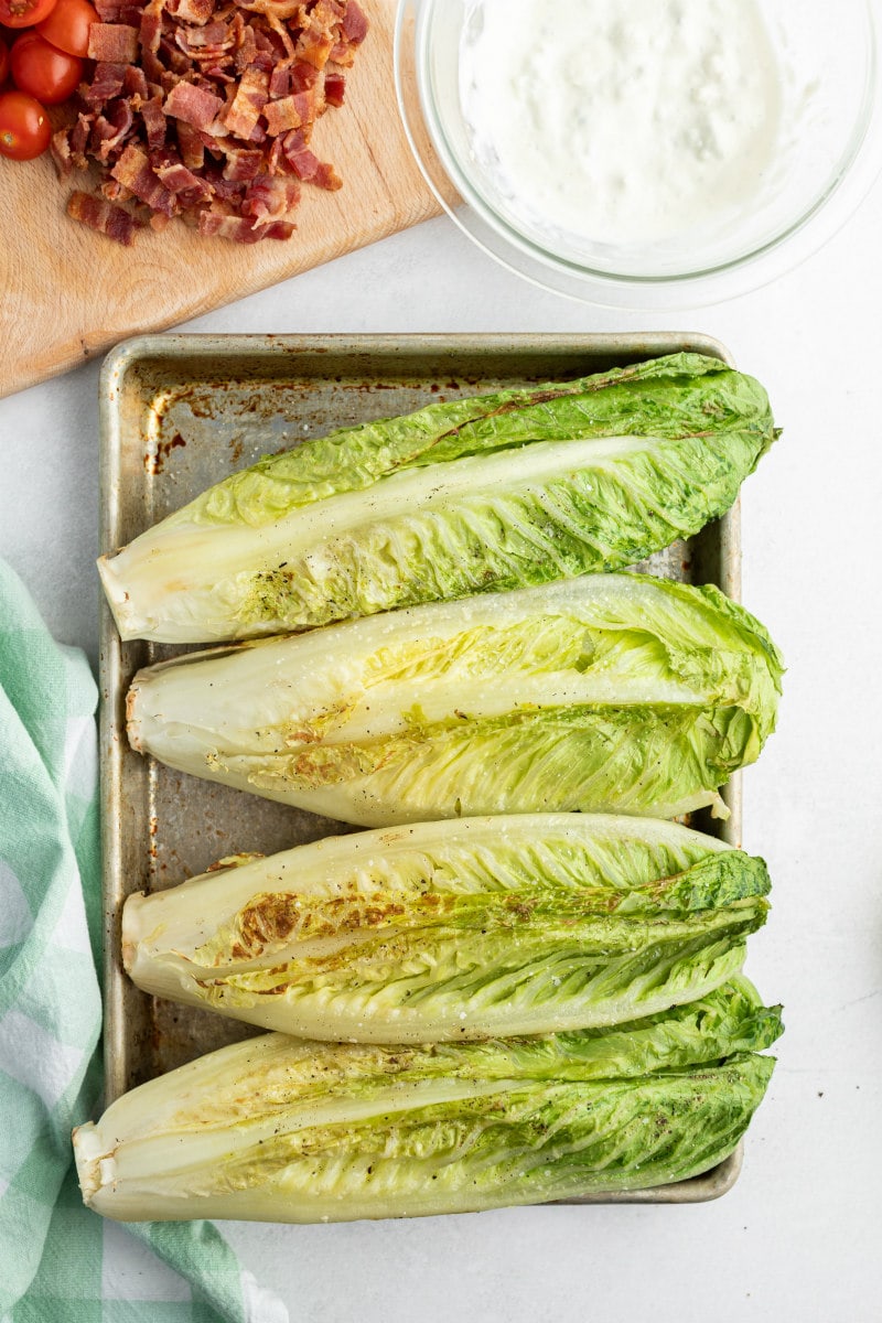 four grilled romaine lettuce heads on a sheet pan with a peek of chopped bacon and blue cheese dressing in the background