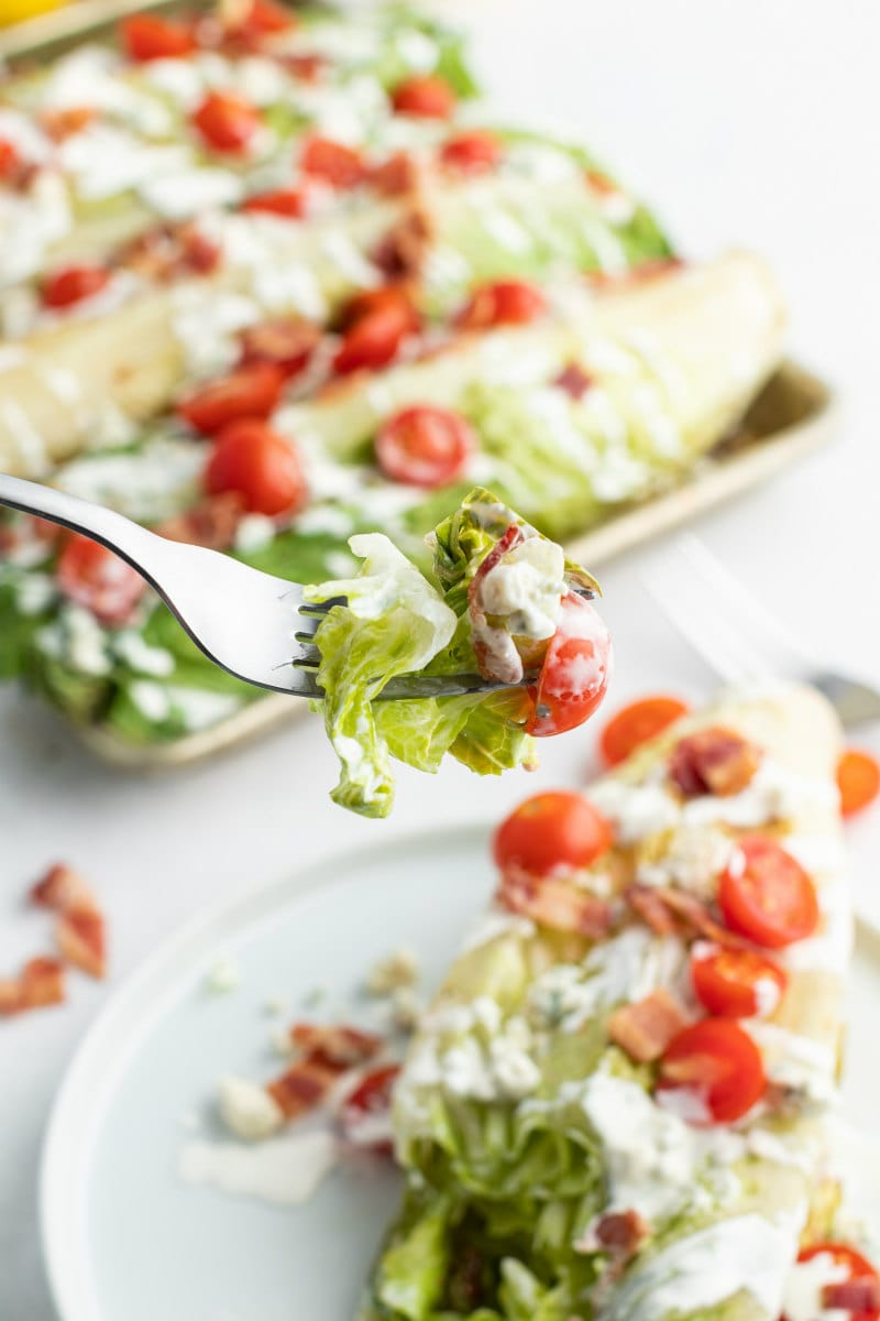 forkful of grilled wedge salad above a white plate of a serving of the salad and more wedge salads in the background