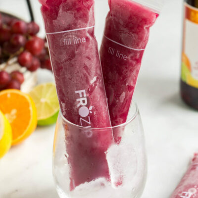 two sangria popsicles standing up in a glass with a bottle of sangria in the background and orange slices, lime slices and grapes in the background too