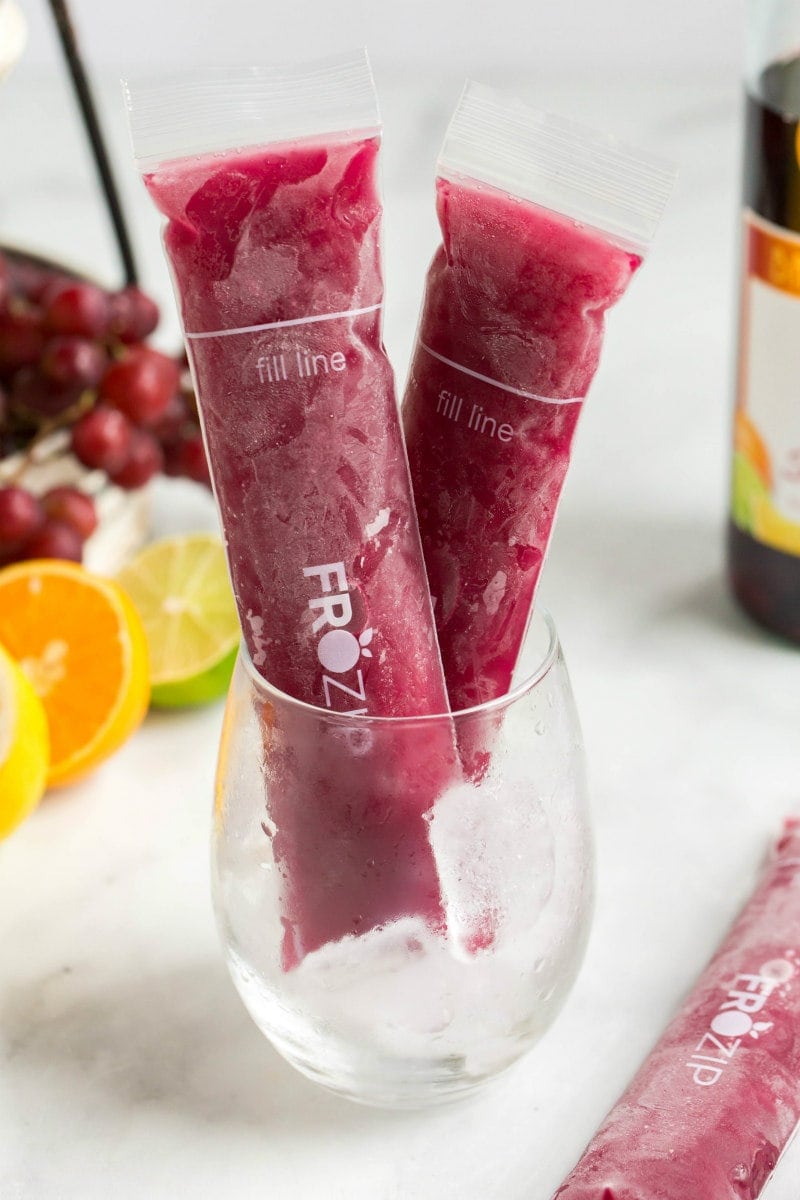 two sangria popsicles standing up in a glass with a bottle of sangria in the background and orange slices, lime slices and grapes in the background too