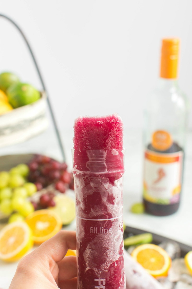 sangria popsicle popping up out of it's wrapper with a hand holding it. fresh fruit and bottle of sangria in background