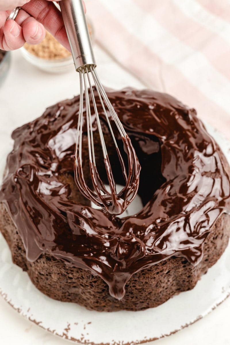 using a whisk to drizzle glaze onto chocolate cake set on a white platter