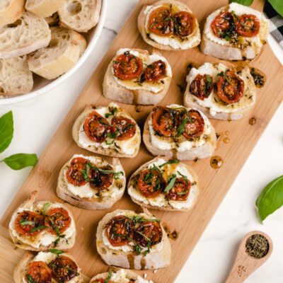 honey tomato bruschetta with ricotta on a wooden board with a bowl of baguette slices and fresh basil on the side with a white and black checked napkin