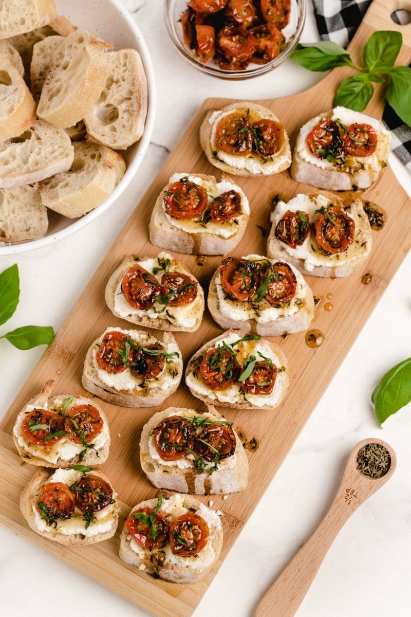 honey tomato bruschetta with ricotta on a wooden board with a bowl of baguette slices and fresh basil on the side with a white and black checked napkin