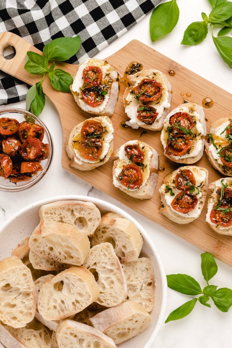 honey tomato bruschetta with ricotta on a wooden board with a bowl of baguette slices and fresh basil on the side with a white and black checked napkin