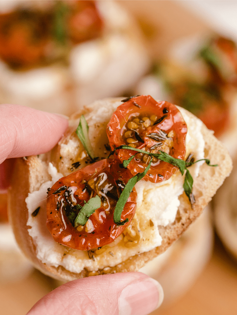 fingers holding a piece of honey tomato bruschetta with ricotta