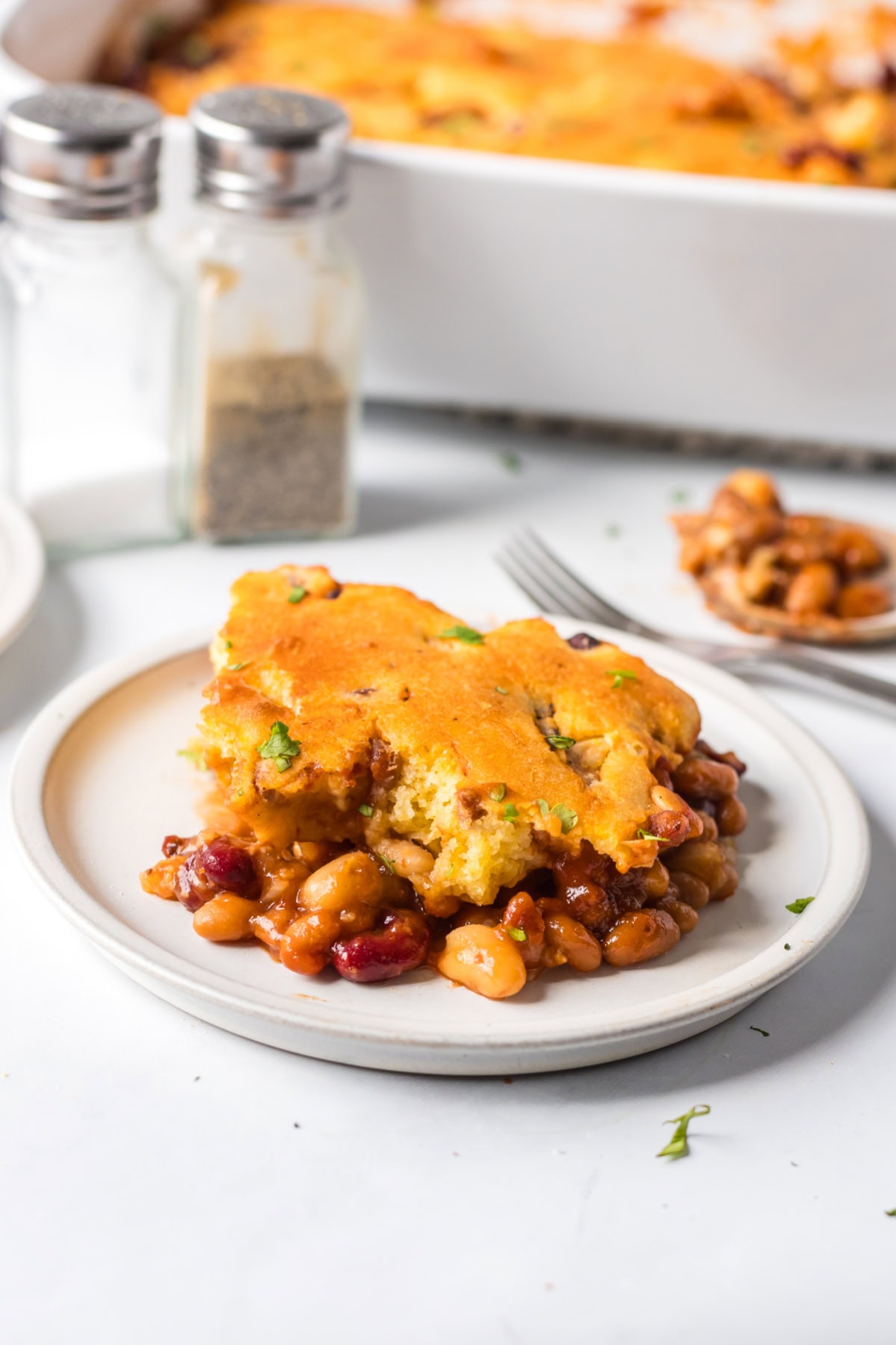 Cowboy Casserole is a hearty, baked beans and cornbread casserole full of smoky, sweet, and savory flavor — perfect for cozy family dinners or potlucks! Recipe from RecipeGirl.com. Serving of beans and cornbread casserole with melted cheese and herbs.