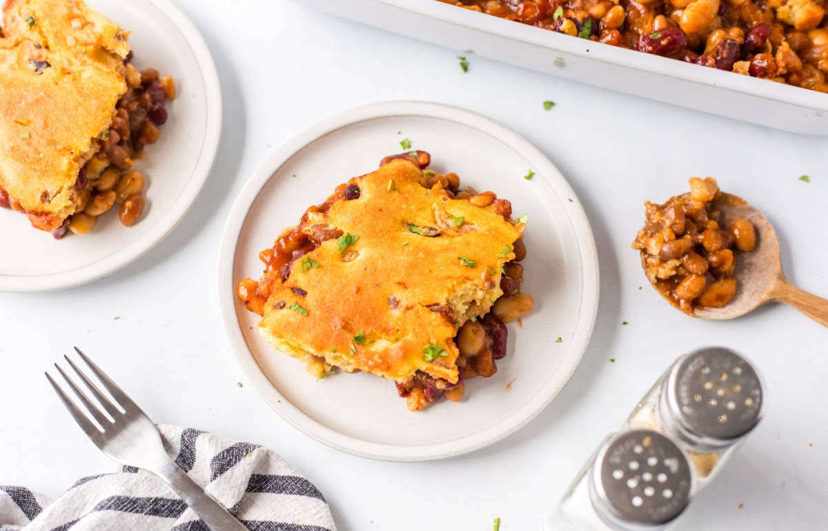 Cowboy Casserole is a hearty, baked beans and cornbread casserole full of smoky, sweet, and savory flavor — perfect for cozy family dinners or potlucks! Recipe from RecipeGirl.com. slice of beans and cornbread casserole on plate
