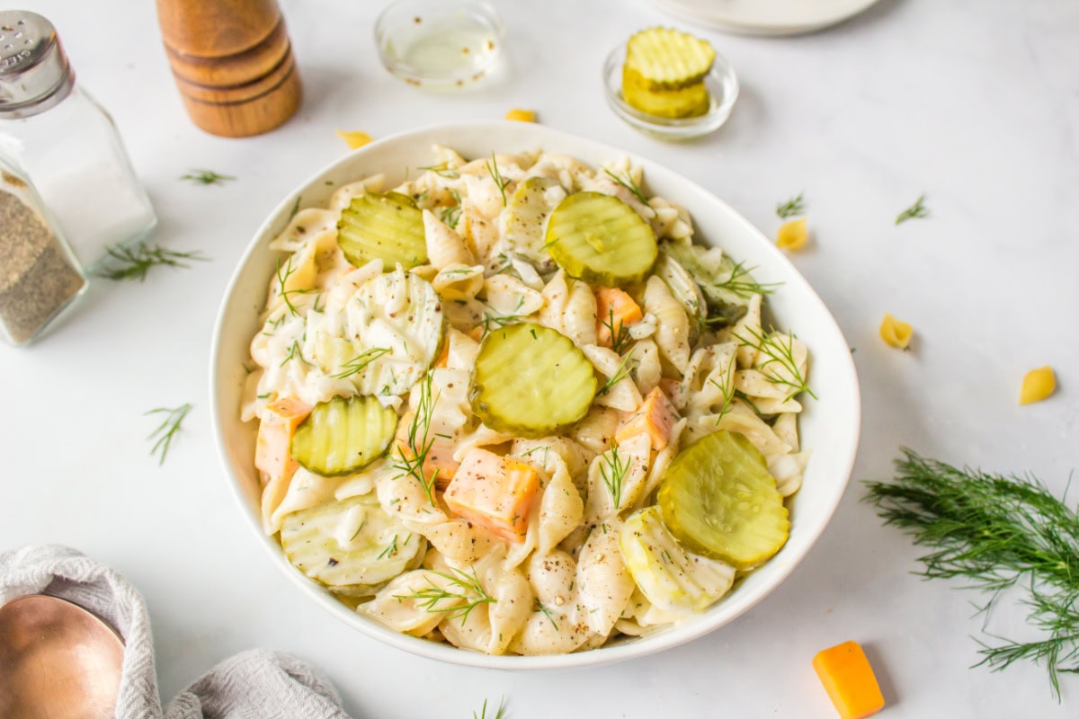 Dill Pickle Pasta Salad is the ultimate creamy, tangy side dish! Shell pasta is soaked in pickle juice, then tossed with cheddar, fresh dill, and a creamy dressing. Perfect for BBQs, potlucks, and summer parties! white bowl with dill pickle pasta salad