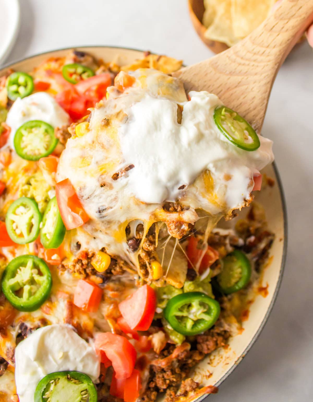 Skillet Nachos 🧀🌶️ The easiest way to make nachos! Tortilla chips topped with seasoned beef, Rotel, black beans, corn, and melty Colby-Jack cheese—add sour cream, guacamole & jalapeños! spoon lifting up serving at skillet nachos