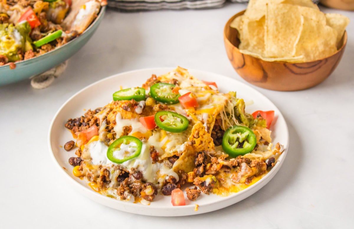 Skillet Nachos 🧀🌶️ The easiest way to make nachos! Tortilla chips topped with seasoned beef, Rotel, black beans, corn, and melty Colby-Jack cheese—add sour cream, guacamole & jalapeños! serving of skillet nachos on a white plate