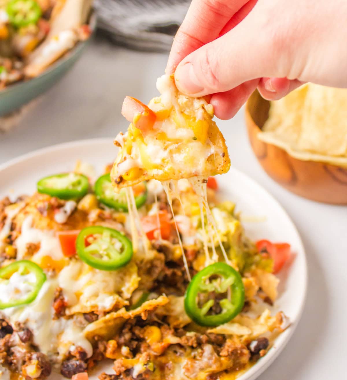 Skillet Nachos 🧀🌶️ The easiest way to make nachos! Tortilla chips topped with seasoned beef, Rotel, black beans, corn, and melty Colby-Jack cheese—add sour cream, guacamole & jalapeños! hand picking up cheesy chip of plate of nachos