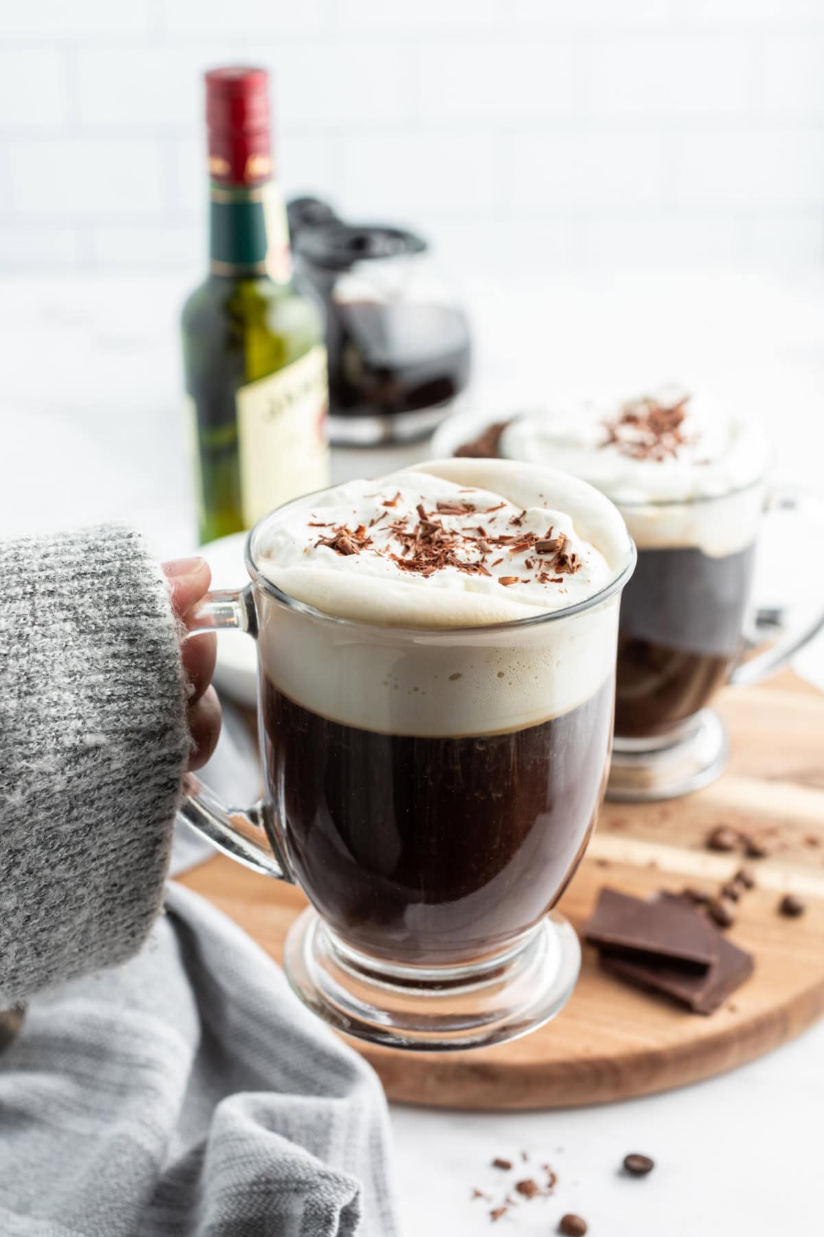 Learn how to make Irish Coffee—hot coffee, Irish whiskey, brown sugar, and whipped cream. A cozy classic cocktail. hand holding cozy mug of irish coffee