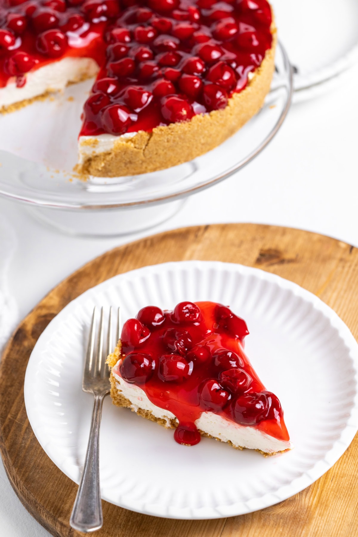 This easy No Bake Cherry Cheesecake has a buttery graham cracker crust, fluffy cream cheese filling, and sweet cherry topping. Perfect make-ahead dessert! slice of no bake cherry cheesecake on plate with rest of cheesecake in background