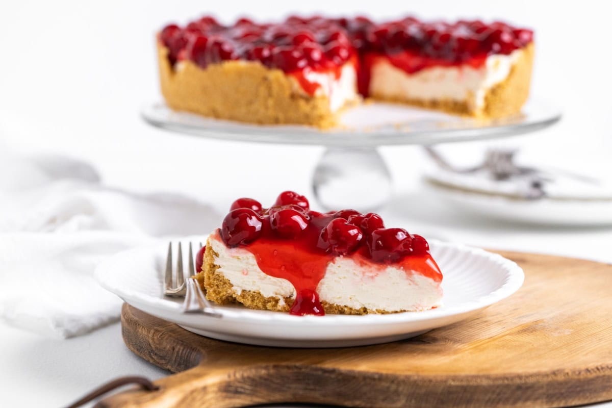 This easy No Bake Cherry Cheesecake has a buttery graham cracker crust, fluffy cream cheese filling, and sweet cherry topping. Perfect make-ahead dessert! slice of no bake cherry cheesecake on plate with rest of cheesecake in background