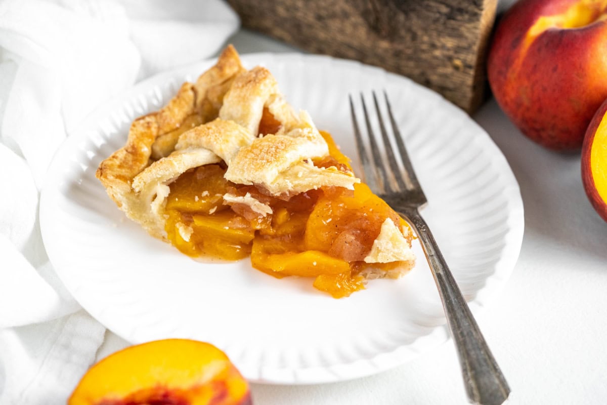 This Fresh Peach Pie Recipe is everything summer baking should be! 🍑 Made with juicy ripe peaches, a flaky homemade butter crust and a gorgeous lattice top, this classic pie is perfect for holidays, BBQs and family gatherings. Save this recipe for peach season! #peachpie #freshpeaches #summerdesserts #pierecipe #homemadepie slice of fresh peach pie on plate