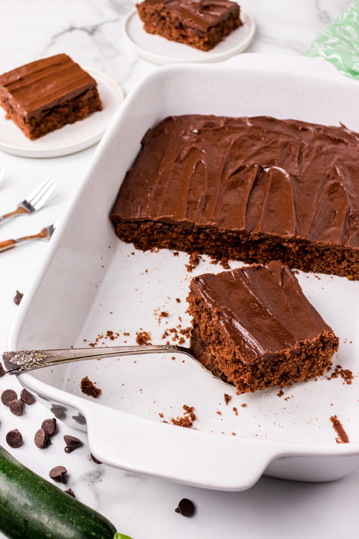 This Chocolate Zucchini Cake is unbelievably moist and rich with chocolate flavor! 🍫 Fresh zucchini keeps the cake soft and tender, while an old-fashioned chocolate marshmallow frosting makes every bite irresistible. Save this easy summer baking recipe! #zucchinicake #chocolatecake #zucchinirecipes #summerbaking #dessertrecipes chocolate zucchini cake in baking pan with slice taken out