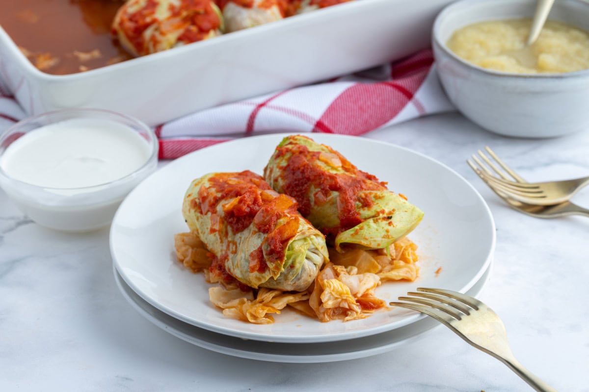 These homemade Polish Cabbage Rolls (Golabki) are the ultimate comfort food! Tender Savoy cabbage leaves are filled with ground beef, rice and seasoned tomato sauce, then baked until tender. Serve with sour cream and applesauce for a delicious old-fashioned family dinner. Save this recipe for a cozy weekend meal! two polish cabbage rolls on a plate